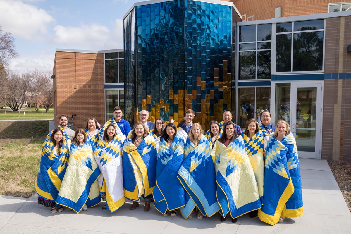 Seventeen men and women wearing yellow, blue and white star quilts pose in two rows with President Barry Dunn in the back row. They are standing on the concrete sidewalk outside of the American Indian Student Center, smiling with plumes and feathers on each head, on a sunny day.