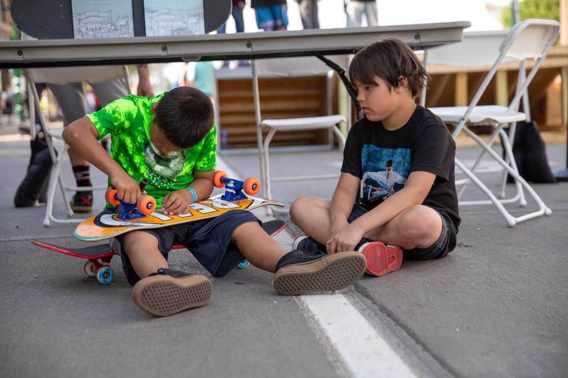 Two children from Pine Ridge sit on the ground together. One is sitting on a skateboard while carefully fixing the skateboard of his companion.