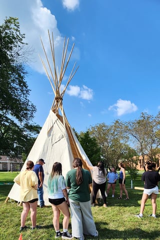 Group of students stand around a newly structured Tipi in the middle of campus grasses