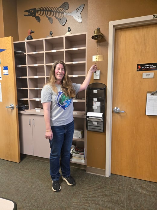 Alison Coulter stands in an office next to a wall-mounted brass bell, holding the bell’s pull chain raised. Behind the person is a cubby-style mail organizer with labeled slots, some containing papers. On top of the organizer are decorative duck figures, and above them is a metal wall art piece shaped like a fish skeleton. To the right is a wooden door with a small sign and a wall-mounted organizer labeled “AFS” holding documents. The walls are brown, and the floor is carpeted.