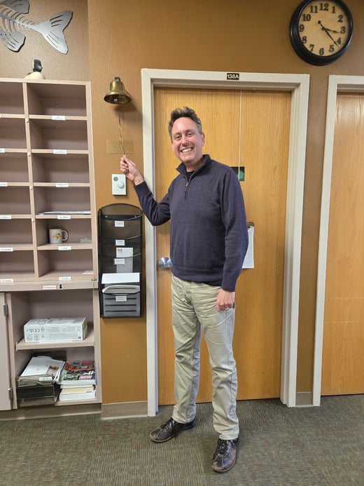 Josh Leffler stands in front of a wooden door labeled “138A,” holding up the pull chain of a wall-mounted brass bell. To the left is a cubby mail organizer with labeled compartments and a few items inside, including a mug and papers. A vertical black wall file organizer labeled “AFS” hangs beside a thermostat. Above the door is a round wall clock. The office has brown walls and carpeted flooring.