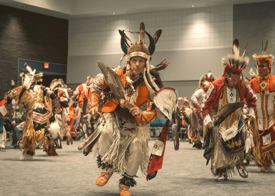 Indigneous dancer in their traditional regalia in center of image while surrounded by other dancers in the arena