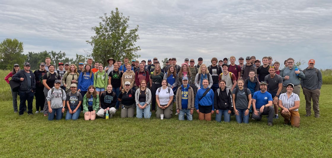 Large group of SDSU NRM students, faculty, and graduate teaching assistants gathered outdoors at Oak Lake Field Station for the NRM 230 Techniques course, standing and kneeling together in a grassy field with trees and a cloudy sky in the background.