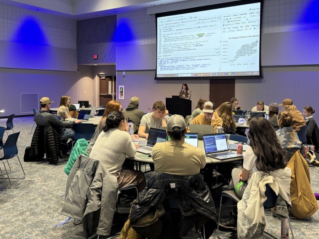 A large annual conference session taking place in a modern, softly lit room where groups of attendees are seated around round tables, each with open laptops and notebooks. In the center foreground, multiple participants face forward while keeping their attention on a presenter standing behind a podium at the front of the room. A large projection screen above the presenter displays lines of code alongside a data visualization chart, reinforcing the technical nature of the talk. Throughout the room, coats are draped over chairs and personal bags rest on the floor, indicating an extended working session. The arrangement of the seating and the orientation of heads and screens suggest that attendees are actively listening and following along with the presenter’s explanation, creating a focused and engaged atmosphere typical of an annual conference workshop or lecture.