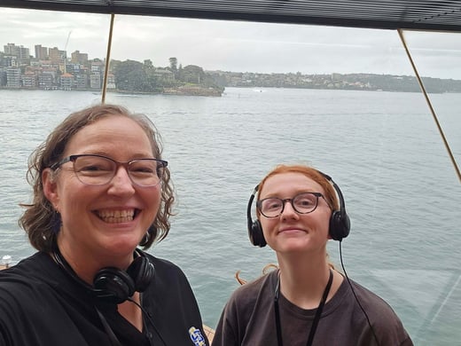Lora Perkins and Chloe Boyda wearing headphones take a selfie on a boat, with water and a shoreline of buildings visible behind them under an overcast sky.
