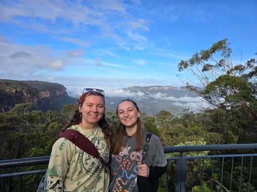 Grace Carruthers and Alaina Wolter stand together at a scenic mountain overlook with a blue sky, clouds, and forested cliffs in the background, posing in front of a railing.