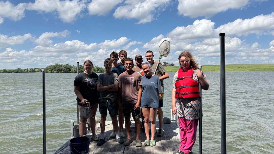 Group of youth campers standing on a dock by Oak Lake during an outdoor field activity, with one participant holding a net and another wearing a life jacket.