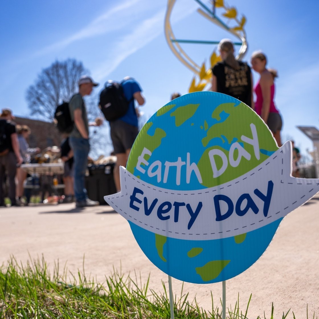 A close-up of a sign shaped like the Earth in the foreground reads “Earth Day Every Day.” In the background, slightly out of focus, a group of people stands outdoors on a sunny day near tables and displays, suggesting an event or gathering. The scene takes place on a campus walkway with grass, trees, and a large decorative sculpture visible, creating an open and vibrant outdoor setting for an Earth Day celebration.