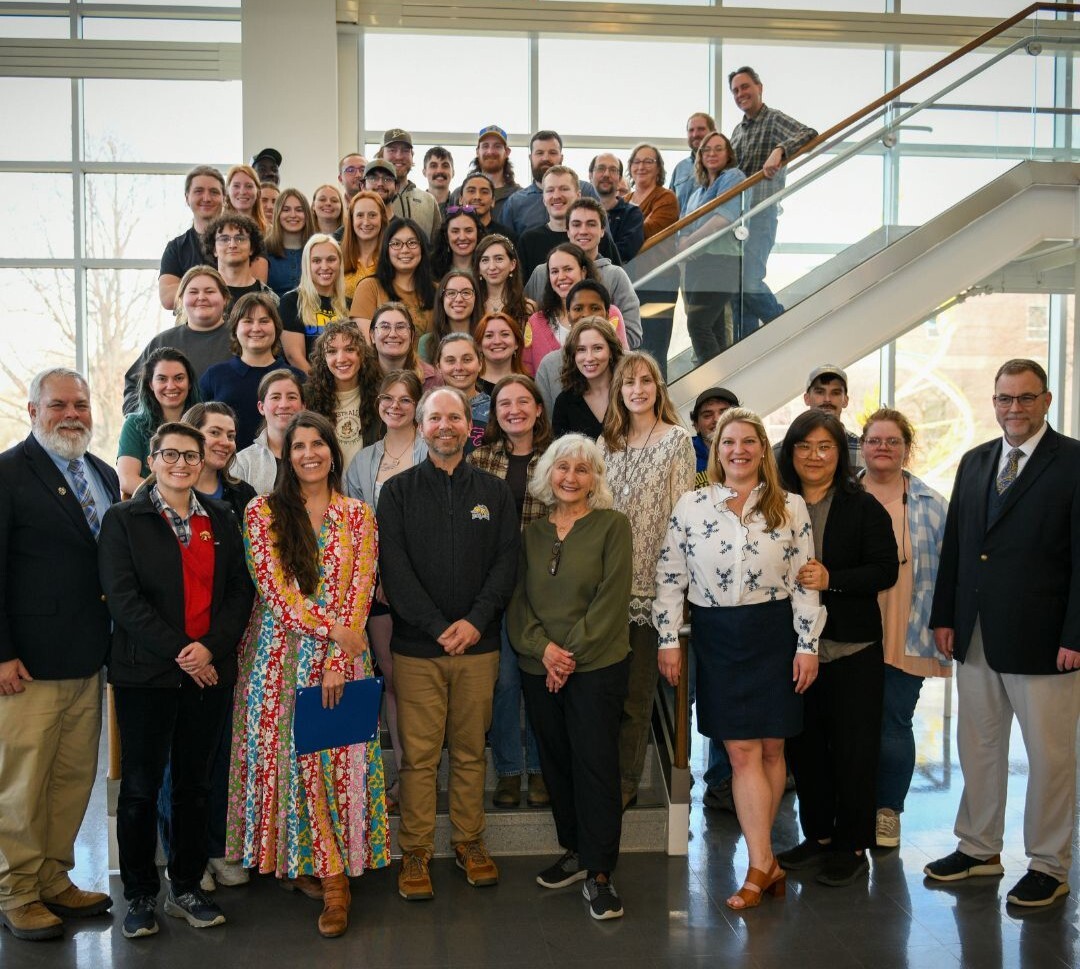 A large group of graduate students and faculty members stands together on a staircase inside a bright building with large windows. The group is arranged in several rows, with individuals standing on steps and along the railing. People are dressed in a mix of casual and semi-formal attire. The background includes glass panels, metal railings, and natural light, creating an open, indoor campus setting. The photo captures a formal group portrait from a graduate student appreciation event.