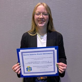 Ava Graham wearing glasses holds a blue-bordered certificate from South Dakota State University recognizing top poster work in Environmental Science and Natural Resource Management.