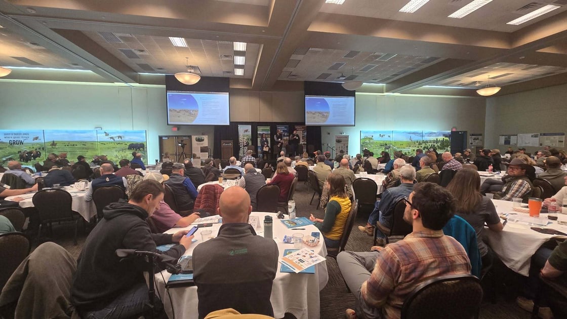 Attendees seated at round tables listen to speakers presenting at the South Dakota Grasslands Summit, with presentation slides displayed on large screens and grassland-themed exhibits and posters along the walls.
