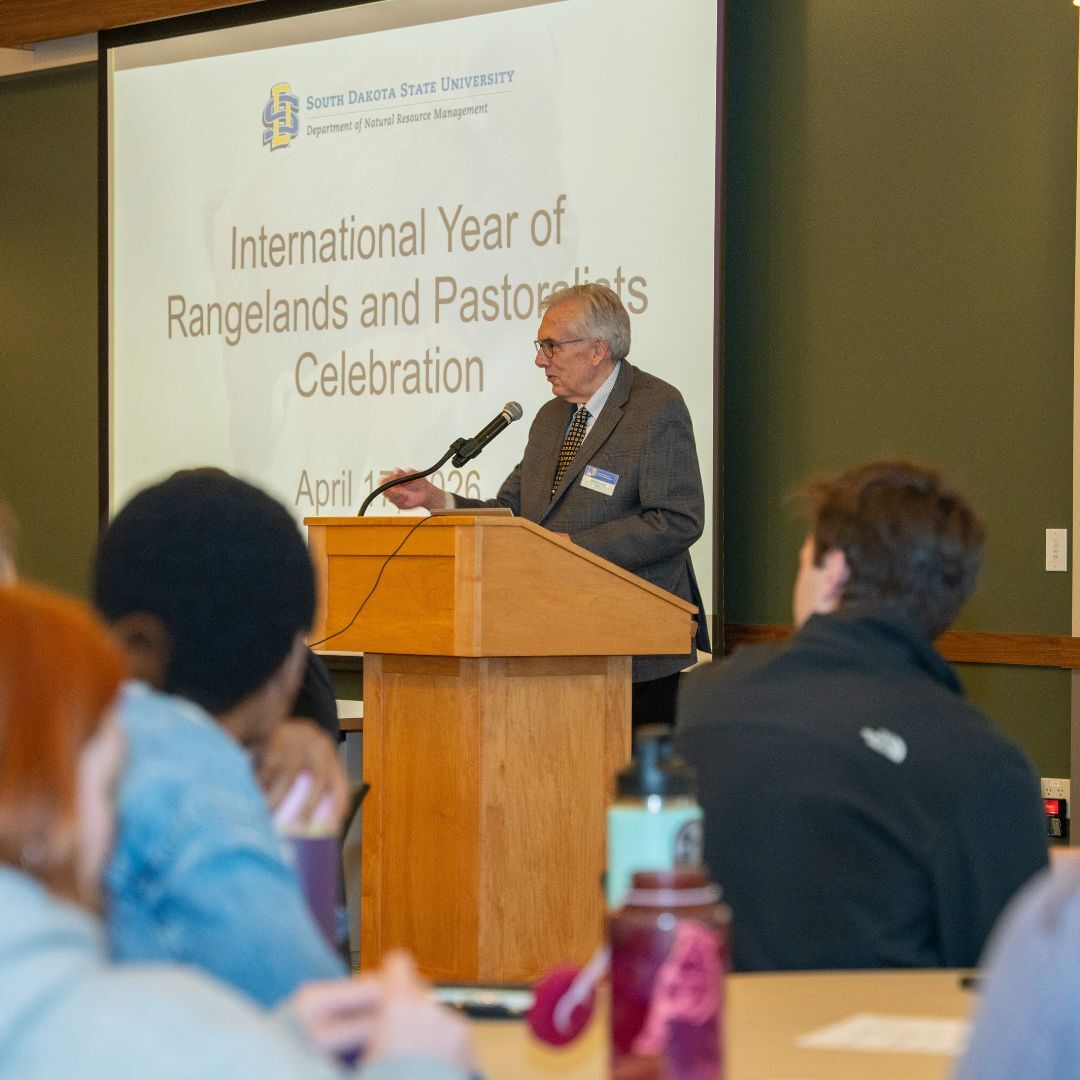 President Dunn stands at a wooden podium speaking into a microphone in the Great hall. Behind him, a projected slide reads “South Dakota State University, Department of Natural Resource Management” and “International Year of Rangelands and Pastoralists Celebration,” with a date in April 2026. An audience sits in the foreground at tables, slightly out of focus, listening to the presentation. The room has green walls, wood trim, and warm indoor lighting.