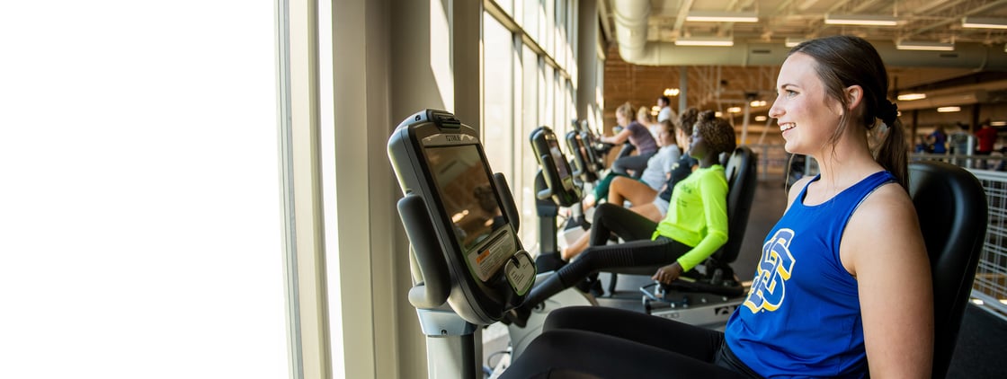 Woman in blue SDSU shirt on exercise equipment in front of a window