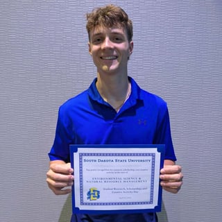 Cooper Olson holds a blue-bordered certificate from South Dakota State University recognizing top poster work in Environmental Science and Natural Resource Management.