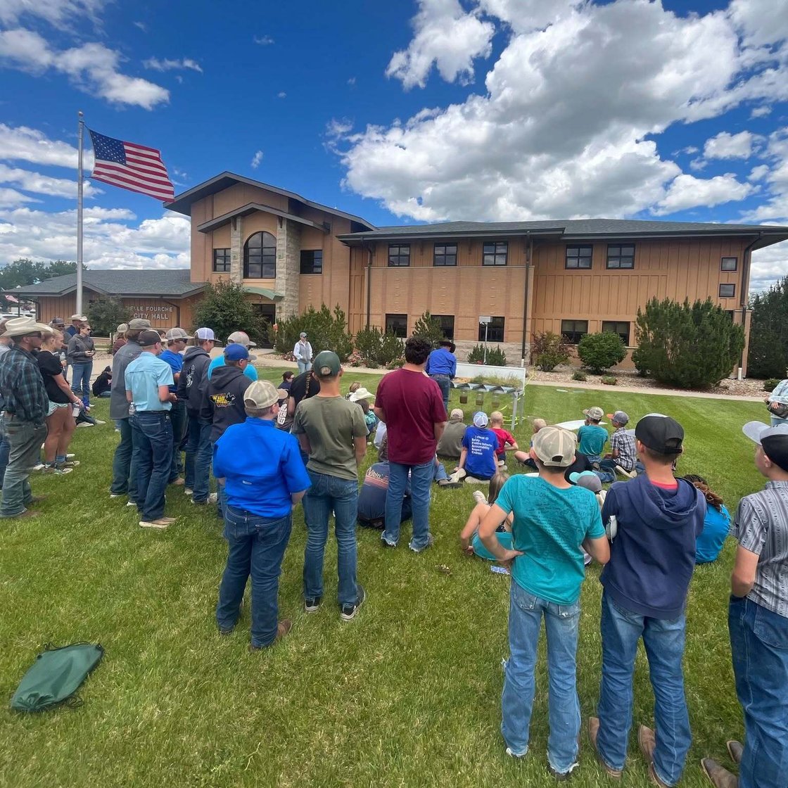 Group of people gathered on a lawn outside a city hall building, with an American flag flying under a blue sky with large clouds.
