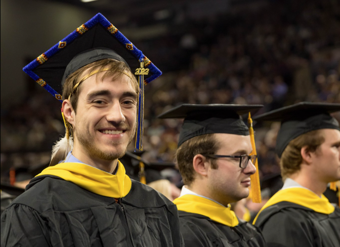 Native American graduate wearing black gown, yellow collar, blue and yellow beaded mortarboard and eagle feather seated next to two other graduates smiles at camera during graduation ceremony. Behind them and out of focus are bleachers filled with onlookers.