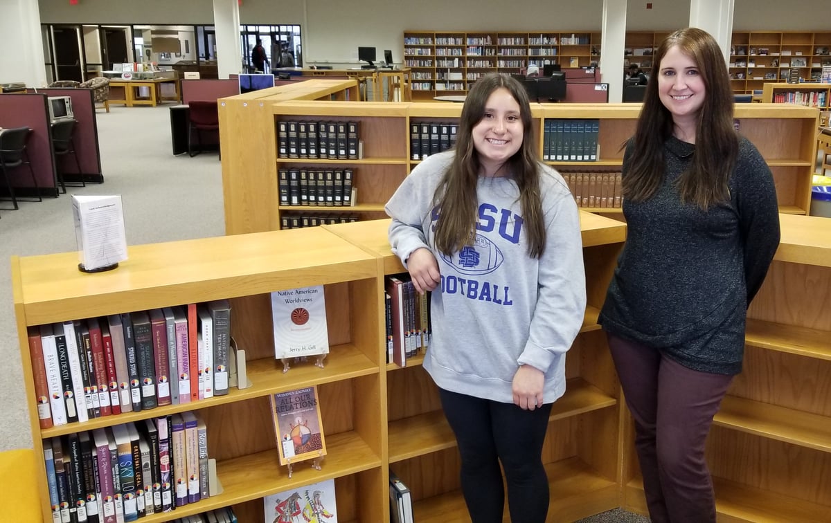 Emirra Returns and Emmeline Weber smiling and posing for a picture inside the Wokini Knowledge Circle in the main level Reading Room of Briggs Library. They are standing among a wall of four-foot high bookshelves full of books on indigenous topics. In the background are computer workstations and more bookshelves.
