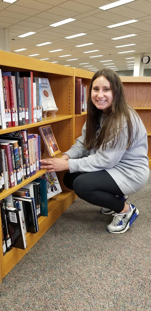 SDSU student Emirra Returns smiling and crouching in front of some low bookshelves, reaching for a book.