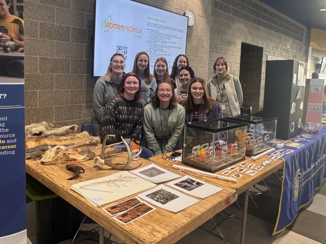 A group of nine graduate students stands behind an informational table at a Women in Science event. The table is covered with natural resource education materials, including animal skulls, antlers, hides, printed plant samples, and two small display tanks with miniature habitat scenes. Additional items such as animal identification cards, photos, and teaching visuals are neatly arranged across the table. Behind the group, a wall-mounted screen displays the “Women in Science” event title and sponsorship information. The indoor setting features brick walls and informational posters, highlighting an interactive exhibit where the students are presenting hands-on learning activities to event participants.