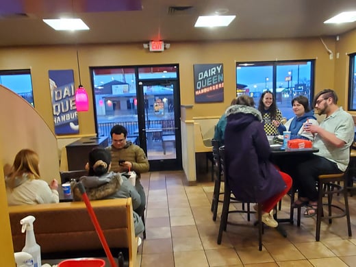 NRM graduate students sit and stand around tables near the entrance, with glass doors and windows visible in the background. Some of them are seated on chairs and stools, while others stand nearby talking. Menu signage is visible further back, along with a wall sign that reads “Dairy Queen.” Daylight is visible through the windows, and the interior features tiled floors and warm lighting.