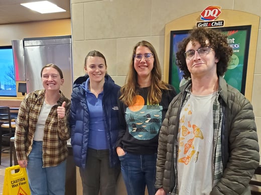 Four graduate students stand close together in front of a wall near the counter area, posing for a group picture. Behind them is a Dairy Queen sign on the wall and part of the service counter. The group is dressed casually. The setting appears clean and well-lit, with neutral-colored walls and restaurant fixtures typical of a fast-food dining area.